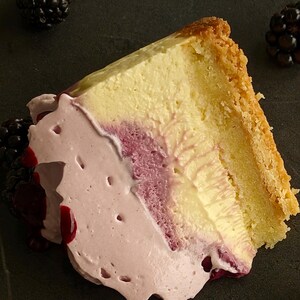 May include: A slice of cheesecake with a light yellow filling and a golden-brown crust. The cake is topped with a pink, whipped cream-like topping and a dark berry sauce. Blackberries are scattered around the cake on a dark surface.
