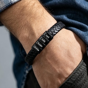 May include: A black braided leather bracelet featuring four rectangular black metal beads. Each bead is engraved with a name: Jack, Julia, Susan, and Emma. The bracelet has a woven texture and a sleek, dark finish, providing a secure fit around the wrist.