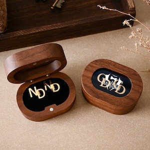 Op de afbeelding: Twee ovale donkerhouten doosjes met magnetische sluitingen. Eén doosje is geopend en toont goudkleurige manchetknopen met de letters ND. Het andere doosje is gesloten en heeft een glazen deksel met een goudkleurig logo van de letters GD en een vlinderdas.