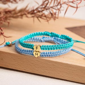 May include: Two macrame friendship bracelets in shades of blue, with gold heart-shaped charms. One bracelet is turquoise, the other is light blue. Each charm has a letter. The bracelets are on a wooden surface.
