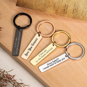 May include: Four metal keyrings with engraved messages. The keyrings are in black, rose gold, gold, and silver. The messages include "I love you to the moon and back", "Love You Forever", and "Drive Safe!!!! I kinda want to keep you".