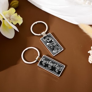 Op de afbeelding: Twee rechthoekige metalen sleutelhangers met een filmstripontwerp. Elke sleutelhanger toont een verticale reeks van drie zwart-witfoto's van een koppel, gevat in een zilverkleurig frame dat is bevestigd aan een ronde metalen ring.