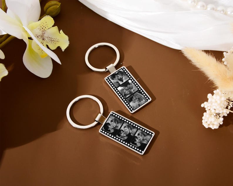 Op de afbeelding: Twee rechthoekige metalen sleutelhangers met een filmstripontwerp. Elke sleutelhanger toont een verticale reeks van drie zwart-witfoto's van een koppel, gevat in een zilverkleurig frame dat is bevestigd aan een ronde metalen ring.