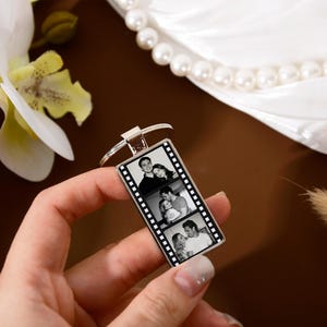 Op de afbeelding: Een rechthoekige zilverkleurige metalen sleutelhanger met een verticaal filmstripontwerp. In het midden van de sleutelhanger staan drie gestapelde zwart-witfoto's van een koppel. De sleutelhanger is bevestigd aan een ronde metalen ring.