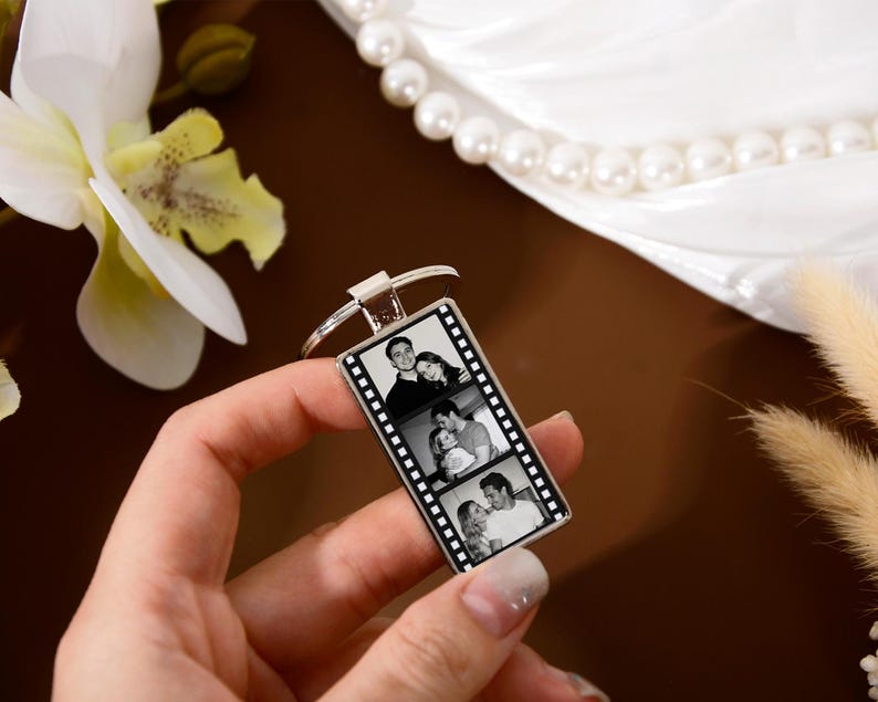 Op de afbeelding: Een rechthoekige zilverkleurige metalen sleutelhanger met een verticaal filmstripontwerp. In het midden van de sleutelhanger staan drie gestapelde zwart-witfoto's van een koppel. De sleutelhanger is bevestigd aan een ronde metalen ring.