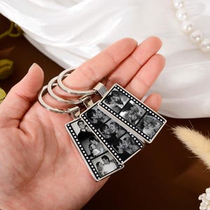 Op de afbeelding: Drie rechthoekige metalen sleutelhangers met een zwart-wit filmstripontwerp. Elke sleutelhanger toont een reeks kleine, verticale portretfoto's, ingesloten in een zilverkleurig frame met een ronde metalen ring eraan bevestigd.