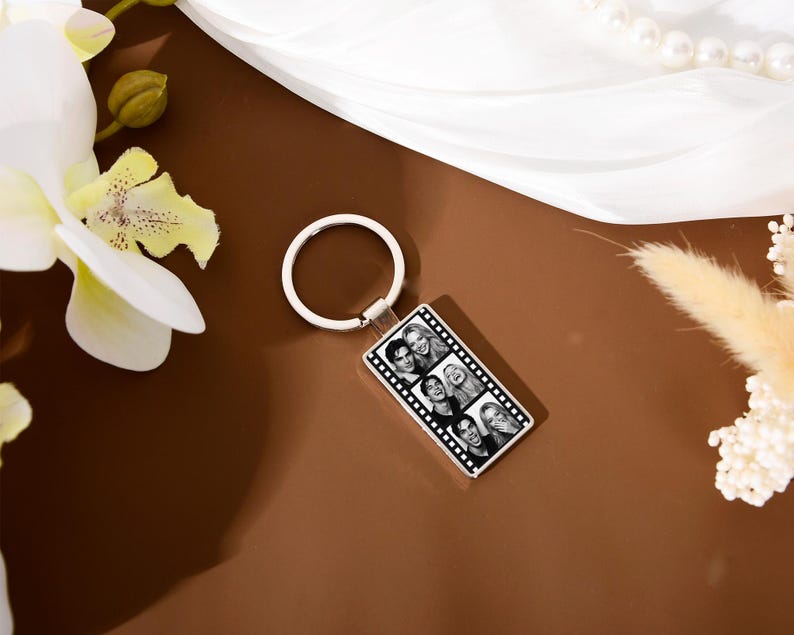 Op de afbeelding: Een rechthoekige metalen sleutelhanger met een zwart-wit filmstripontwerp. Het midden van de sleutelhanger toont een verticale reeks van vier kleine portretfoto's, omlijst door een klassieke filmrand met perforatiegaatjes.