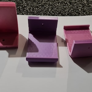 May include: Three 3D printed wall mounts, one purple and two pink. The mounts are designed to hold a phone or other device.