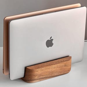 Puede incluir: Un soporte horizontal de madera para portátiles con una base curva que sostiene dos portátiles cerrados en posición vertical. Un portátil es plateado con el logotipo de Apple visible y el segundo portátil detrás es de color oro rosa metálico.
