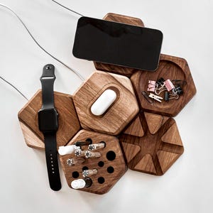 May include: A wooden desk organizer with a hexagonal design. It holds a black smartphone, a black smartwatch, white earbuds, pens, and paper clips. The organizer is made of dark wood and has a modern aesthetic.