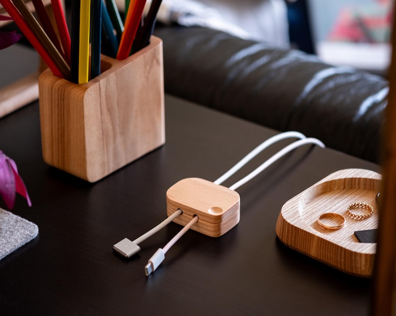 a wooden desk with various items on it, including a wooden pencil holder filled with colored pencils, a wooden tray with a pair of gold rings, and a wooden box with a USB cable plugged into it.
