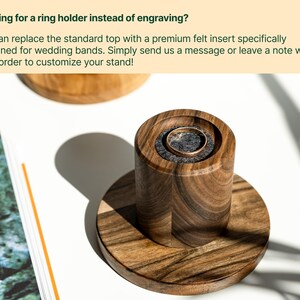 a wooden ring holder with a gold ring inside, placed on a wooden stand. The stand has a textured finish, and the ring holder appears to be made of wood. The background is white, and there is a green plant visible.