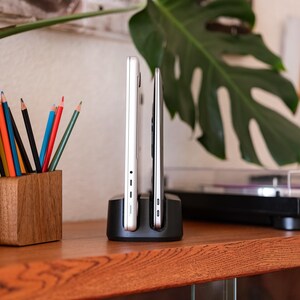 Op de afbeelding: Een zwart oplaadstation houdt twee zilveren elektronische apparaten vast. Een houten potloodhouder met kleurrijke potloden staat links. Een groot groen blad en een platenspeler staan op de achtergrond. De items staan op een houten oppervlak.