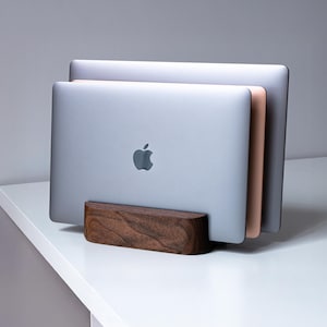Peut inclure: Un support vertical pour ordinateur portable en bois brun foncé contenant deux ordinateurs portables argentés. Le support présente un design en bloc rectangulaire lisse avec une fente centrale qui maintient les appareils en position verticale.