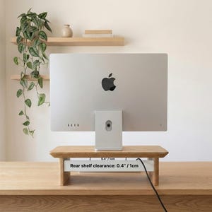 a silver Apple iMac computer monitor placed on a wooden stand, with a potted plant and a shelf with books and a vase on display in the background.