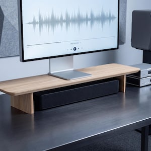a modern, minimalist workspace with a large flat-screen monitor displaying a sound waveform, a wooden desk with a black speaker system, and a gray speaker system.