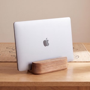 Op de afbeelding: Een zilveren laptop met een zichtbaar Apple-logo staat verticaal in een massief houten standaard. De standaard heeft een glad, gebogen ontwerp met een natuurlijke houtnerfafwerking en biedt een stabiele basis voor het apparaat.