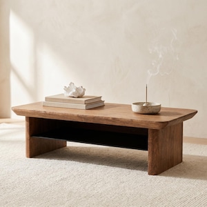 May include: A rectangular wooden coffee table with a lower shelf. On the table are two books, a white coral sculpture, and a small speckled bowl with an incense stick emitting smoke. The table is made of light brown wood.