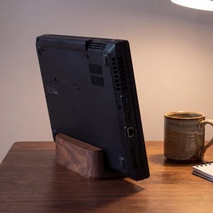 Könnte beinhalten: Ein schwarzer Laptop steht vertikal in einem dunkelbraunen Holzständer. Das Gerät zeigt die Rückseite mit Lüftungsschlitzen sowie verschiedene Anschlüsse, darunter USB und Ethernet, entlang der seitlichen Kante des Laptops.