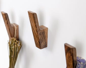 May include: Three wall-mounted coat hooks crafted from dark brown wood with a visible natural grain. Each hook features an angled, upward-pointing design. One hook holds a bundle of dried grass, and another holds a folded purple patterned fabric.