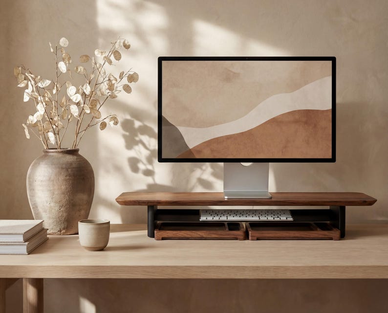 a wooden desk with a computer monitor, keyboard, and a vase with white flowers. The desk is set against a beige wall, and there is a cup and a book on the desk as well.