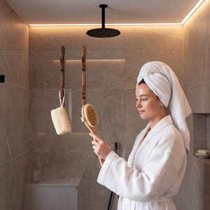 A woman in a white bathrobe stands in a modern bathroom, holding a wooden brush and looking at a mirror.