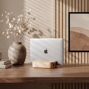 May include: A silver laptop with an Apple logo is held upright in a light-colored wooden stand. The setup includes a ceramic mug, two stacked books, and a vase filled with dried silver dollar eucalyptus branches on a wooden desk.
