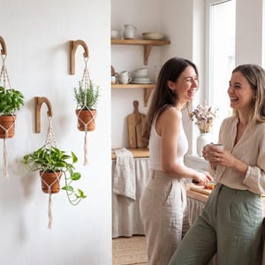 Two women are standing in a kitchen, smiling and holding cups, surrounded by potted plants and kitchenware.