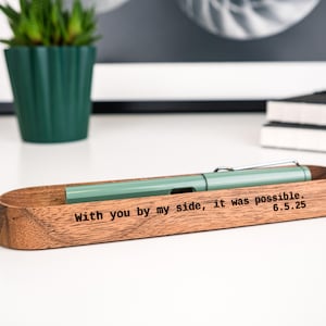 Op de afbeelding: Een houten pennenhouder met een lichtgroene pen erin. De houder heeft de inscriptie "With you by my side, it was possible. 6.5.25" gegraveerd op de voorkant. De pennenhouder is gemaakt van hout en heeft een natuurlijke afwerking.