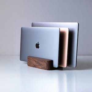 Pode incluir: Um suporte vertical para portátil em madeira castanha escura que segura três portáteis de várias cores, incluindo prateado e ouro rosa, com um logótipo da Apple visível no dispositivo frontal. O suporte apresenta uma base retangular arredondada.