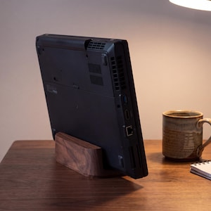 May include: A black laptop computer is positioned vertically in a dark brown wooden stand. The laptop shows its rear panel with ventilation grilles and various ports, including USB and Ethernet, while resting on a matching dark wood desk surface.