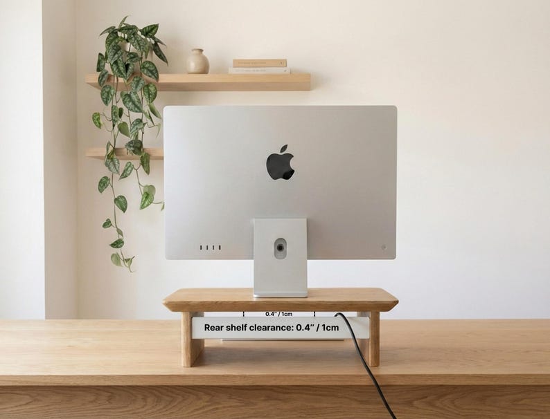 a silver Apple iMac computer monitor placed on a wooden stand, with a potted plant and a shelf with books and a vase on display in the background.