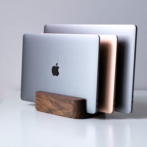 Könnte beinhalten: Ein vertikaler Laptopständer aus dunkelbraunem Holz, der drei Laptops in einer Reihe hält. Der vordere Laptop ist silber mit einem schwarzen Apple-Logo, der mittlere ist roségold und der hintere ist silber. Der Ständer hat eine abgerundete Form.