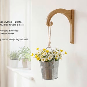 A hanging metal bucket filled with yellow and white flowers is mounted on a wall, with a wooden bracket attached to the wall.