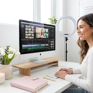 A woman is sitting at a desk, working on a computer with a large monitor, keyboard, and mouse. She is smiling and appears to be focused on her work. The desk is decorated with a vase of flowers, a candle, and a pink notebook.