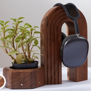 May include: A wooden headphone stand and planter. The dark brown stand features an arched design with grooves and holds dark grey headphones. A small green plant in a black pot sits in the wooden planter base.