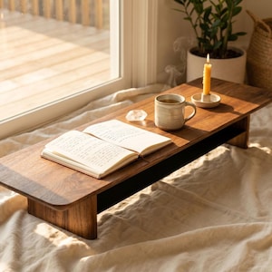 A wooden table with an open book, a mug of coffee, and a lit candle on it, placed near a window.