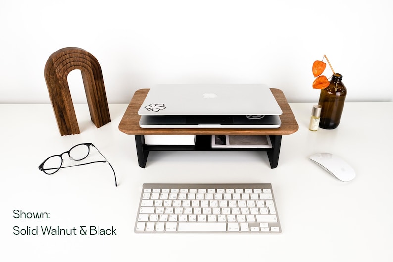a workspace with a laptop, glasses, a keyboard, and a mouse on a white desk. There is also a wooden stand and a vase with orange flowers.
