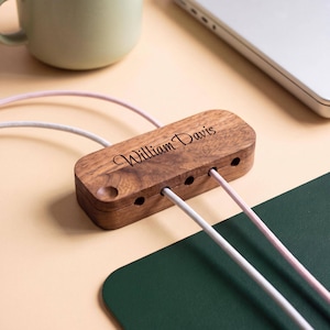 May include: A rectangular wooden cable organiser with a dark brown wood grain finish. The top surface is engraved with the name William Davis in a cursive script. It features five circular cutouts for routing cables, with two light-coloured cords.