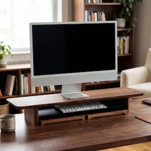 a modern workspace with a wooden desk, a white computer monitor, a keyboard, and a mouse. The desk has a wooden stand that elevates the monitor, providing a comfortable viewing angle. The room also includes a bookshelf filled with books and a plant, creating a cozy and functional environment.