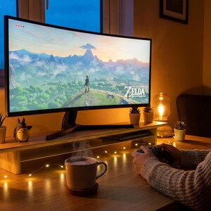 A woman is sitting at a desk, playing a video game on a large monitor. The monitor displays a scenic landscape from the video game, and the woman is holding a video game controller. The desk is decorated with various items, including a mug, a candle, and several potted plants.