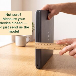 A person's hands are holding a ruler and measuring the size of a closed laptop computer on a wooden desk.
