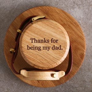 A wooden watch stand with a watch attached to it, and a message engraved on the watch stand that says "Thanks for being my dad."