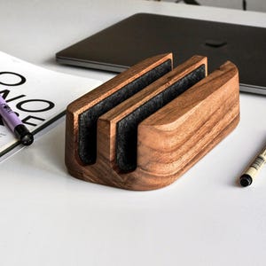May include: A rectangular wooden laptop stand featuring a natural brown wood grain finish and two vertical slots lined with dark gray felt material. The stand is designed to hold a laptop in a vertical position, providing a stable base for desk organization.