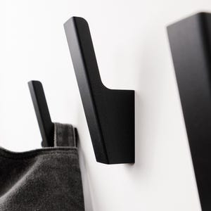Op de afbeelding: Een set van drie matzwarte wandhaken met een hoekig, geometrisch ontwerp. Eén haak houdt een donkergrijs stoffen item vast, terwijl de andere twee haken leeg zijn en op een effen wit oppervlak zijn gemonteerd, wat een strak en modern profiel toont.