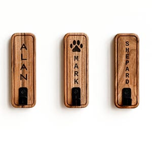 Op de afbeelding: Drie rechthoekige houten wandhaken met afgeronde hoeken. Elke haak heeft onderaan een zwarte metalen bevestiging. De linkerhaak is gegraveerd met de naam ALAN, de middelste met een pootafdruk en MARK, en de rechterhaak met SHEPARD.