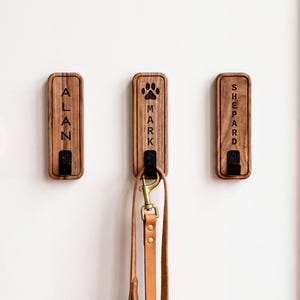 Könnte beinhalten: Drei rechteckige Wandhaken aus Holz mit abgerundeten Kanten, jeweils mit einem schwarzen Metallhaken ausgestattet. Der linke Haken ist mit dem Namen ALAN graviert, der mittlere mit einer Pfote und MARK, der rechte mit SHEPARD. Eine Lederleine hängt am mittleren Haken.