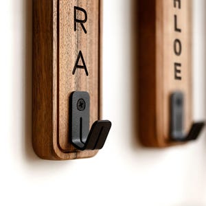 May include: Vertical rectangular wooden wall hooks featuring a natural brown wood grain finish. Each hook includes a black metal bracket secured with a screw. The wood displays black vertical lettering with the visible characters R, A, and L, O, E.