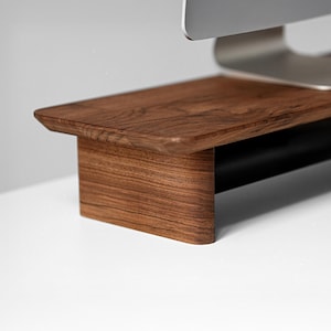 Puede incluir: Un soporte de monitor de madera marrón oscuro con una superficie lisa y pulida y bordes redondeados. El soporte cuenta con una plataforma superior horizontal apoyada por dos patas laterales curvas para una base elevada.