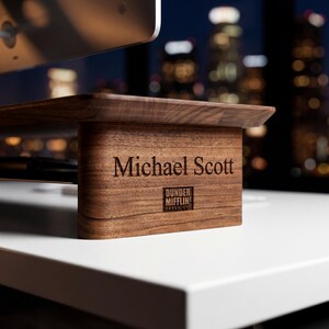 May include: A wooden monitor stand with the name "Michael Scott" engraved on the front. The stand is made of dark wood and has the "Dunder Mifflin Paper Co." logo. The stand is on a white surface.
